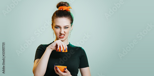 teenager girl grimacing with halves an orange on a studio green background, impudent character and behavior, rebel girl woman raised lip edge showing teeth and squeezes juice from fruit