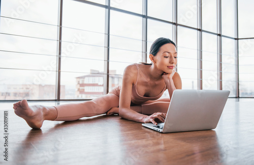 Brunette with slim body type doing exercises on the floor and using laptop