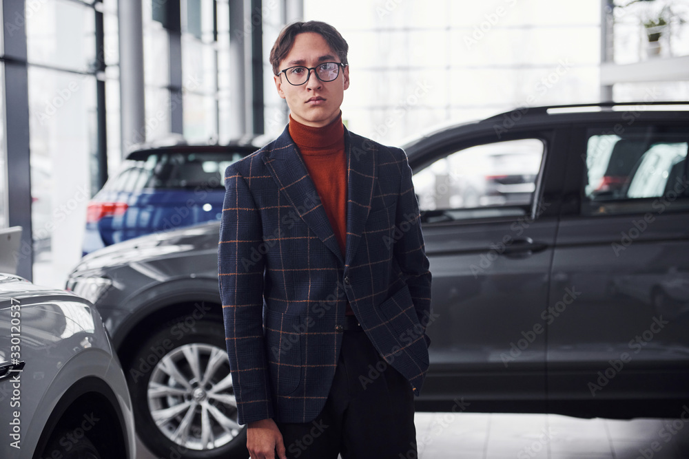Posing for the camera. Young business man in luxury suit and formal clothes is indoors near the car Posing for the camera. Young business man in luxury suit and formal clothes is indoors near the car