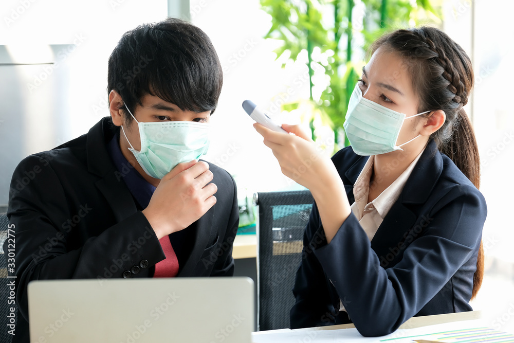 Women in the office use thermoscan to check temperature of the man who wear black suit. There are notebook, chart, pen and pencil on the table. Protection virus and business concept.