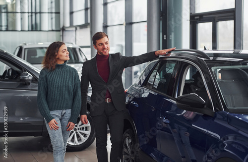 Professional salesman assisting young girl by choosing new modern automobile indoors