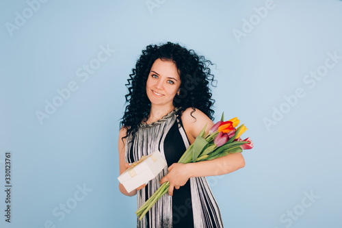 Young beautiful girl with curly hair holding a box and a bouquet of flowers in hands on a blue background