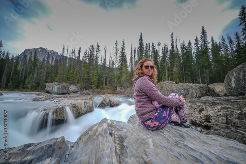 Obraz na płótnie Young woman is sitting on a rock, in the back a small waterfall in a forest, long exposure, Mount Revelstoke National Park bij Revelstoke w salonie