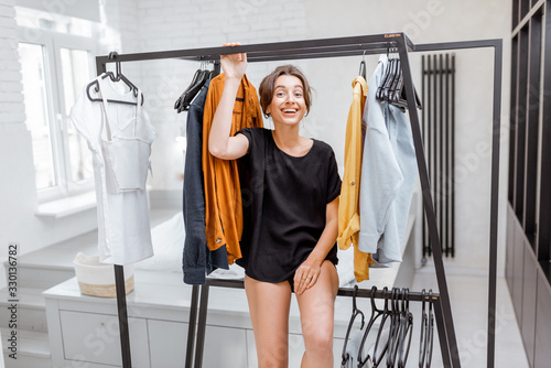Portrait of a young and cheerful woman choosing casual clothes to wear, standing between hangers and looking out of the clothes in the wardrobe at the bedroom