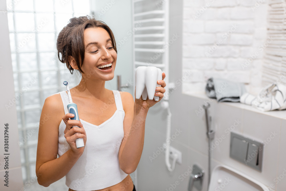 Portrait of a young and cheerful woman standing with electric toothbrush during morning hygiene procedures in the bathroom Portrait of a young and cheerful woman standing with electric toothbrush during morning hygiene procedures in the bathroom