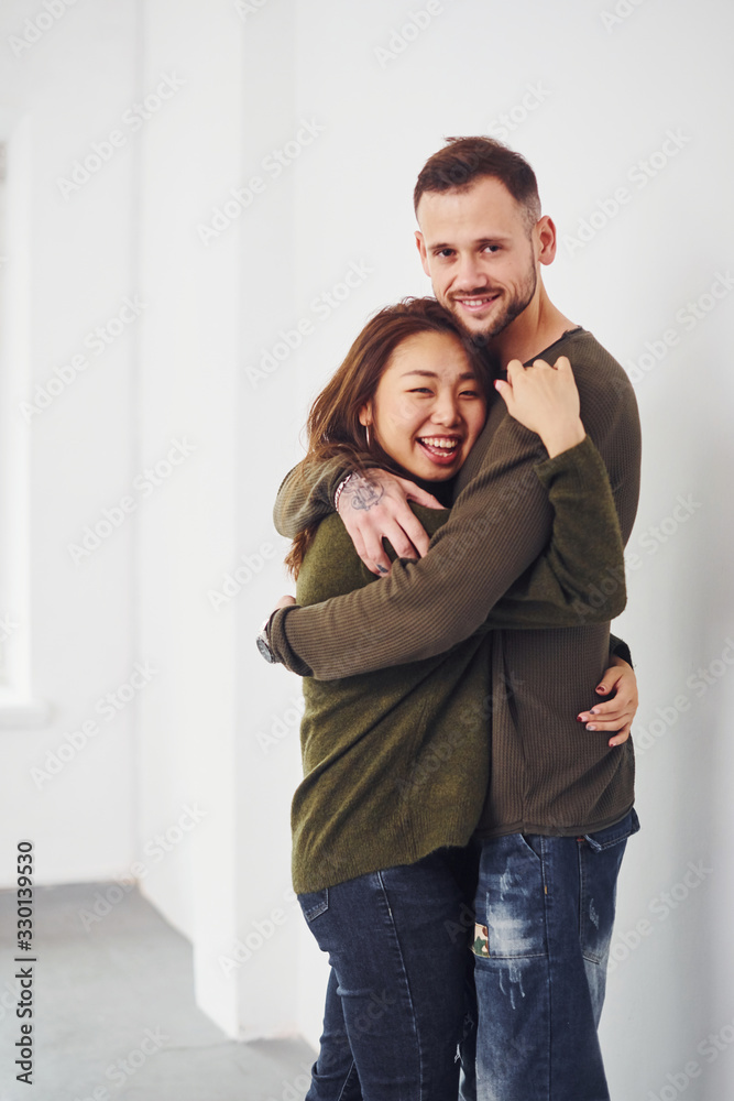 Happy multi ethnic couple in casual clothes embracing each other indoors in the studio. Caucasian guy with asian girlfriend