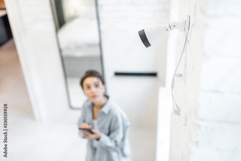 Woman controls home video surveillance with a smartphone indoors, view from the above and ip camera on the foreground. Concept of a wireless home security systems Woman controls home video surveillance with a smartphone indoors, view from the above and ip camera on the foreground. Concept of a wireless home security systems