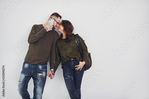 Cheerful multi ethnic couple with backpack and phone standing together indoors in the studio against white background