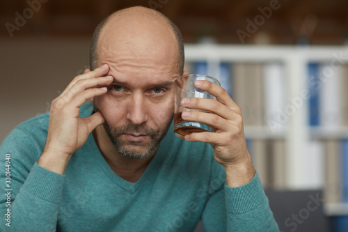 Portrait of drunk mature man holding glass with whiskey and looking at camera with sad sight
