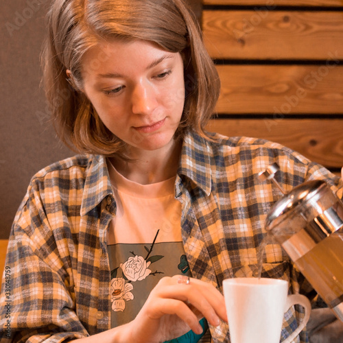 A blonde girl in a plaid shirt at a table in a cafe and drinks tea or coffee.