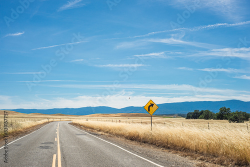 A rural road with a yellow curve sign in the Northern California countryside, with mountains in the background