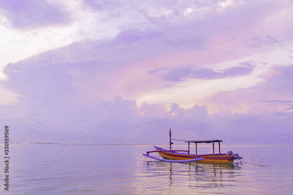 Traditional Balinese wooden fishing boat at dawn on Sanur beach in Bali. Cloudy pink dawn sky. Reflection in the water. Calm at sea