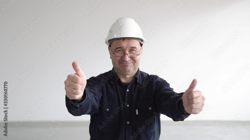 Man, aged, in white construction, engineering helmet, in blue shirt white buttons. With glasses. Showing approving thumbs up both hands and smiling camera. White room. Ability insert text graphics Man, aged, in white construction, engineering helmet, in blue shirt white buttons. With glasses. Showing approving thumbs up both hands and smiling camera. White room. Ability insert text graphics
