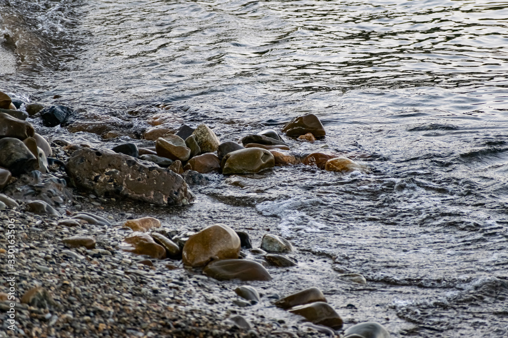 Obraz premium Large and smooth stones in the coastal waves of salty sea water. The coast of the black sea foams and roars, beating against a huge pebble, illuminated by sunlight