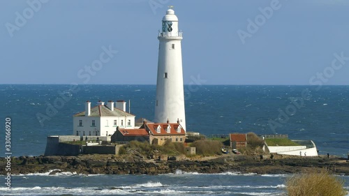 Sunlit lighthouse and properties against a blue sky, with unidentifiable visitors walking around. A tidal causeway links St Mary’s Lighthouse back to the nearby shore at Whitley Bay, England, UK. photo