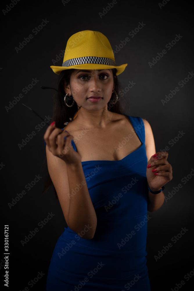 fashion portrait of an young and attractive Indian Bengali brunette girl with blue western dress, yellow cap and sunglass in front of a black studio background. Indian fashion portrait and lifestyle. fashion portrait of an young and attractive Indian Bengali brunette girl with blue western dress, yellow cap and sunglass in front of a black studio background. Indian fashion portrait and lifestyle.