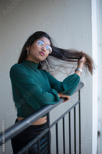 Fashion portrait of an young and attractive Indian Bengali brunette girl with green cropped top, skirt and spectacles standing on a balcony in white background. Indian fashion portrait and lifestyle.