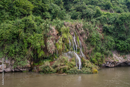 Guilin, China - May 10, 2010: Along Li River. Waterfall off karst mountain under silver sky with multiple silver-white streams of water anong green and brown foliage. Lands in brown water. photo