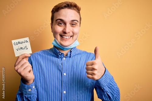 Young man wearing medical security mas holding paper note reminder with coronavirus alert word happy with big smile doing ok sign, thumb up with fingers, excellent sign photo