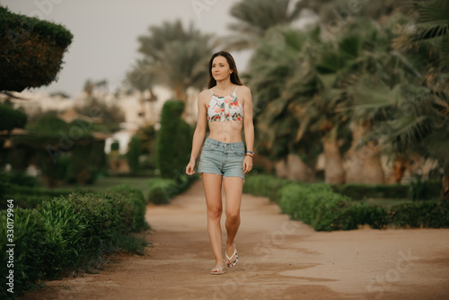 A young model walking on the sand path in the garden with palm trees in a swim top and denim shorts near the red sea in the morning
