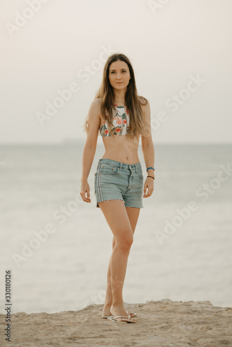 An attractive model with long hair relaxing on the rocky shore of the red sea  in a swim top and denim shorts on the sunrise