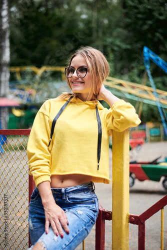 Young blond woman, wearing yellow hoody, blue jeans, spending time in amusement theme park in summer. Three-quarter portrait of pretty girl, leaning on fence railing in front of colorful ferris wheel