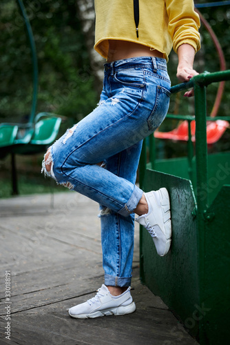 Close-up picture of legs of young woman, wearing yellow hoody, blue jeans, standing on old carrousel leaning on green railing in park in summer © Natalia