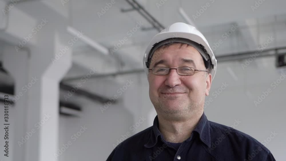 An adult, a credible engineer, an architect in a blue shirt, stands in a large white room with columns, in a white protective helmet. He puts on glasses for vision and smiles at the camera An adult, a credible engineer, an architect in a blue shirt, stands in a large white room with columns, in a white protective helmet. He puts on glasses for vision and smiles at the camera
