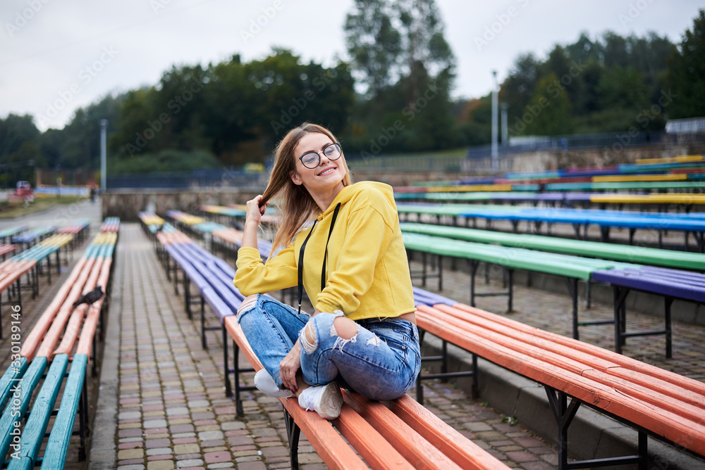 Young blond woman, wearing yellow hoody, blue jeans and eyeglasses, sitting on colorful benche in city urban park in summer. Portrait of pretty girl, relaxing resting on sunday afternoon. Leisure time