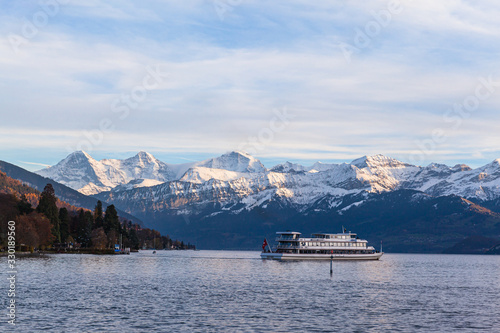 Stunning panorama view of famous Swiss Alps peaks on Bernese Oberland Eiger North Face, Monch, Jungfrau at dusk from Lake Thun (Thunersee) on sunny autumn day with cruise ship, Bern, Switzerland