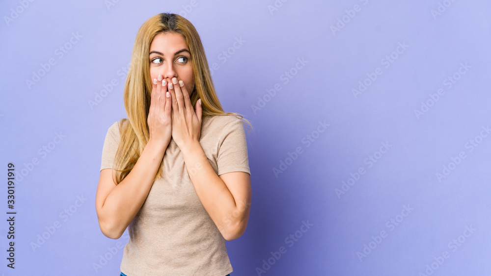 Young blonde woman isolated on purple background thoughtful looking to a copy space covering mouth with hand. Young blonde woman isolated on purple background thoughtful looking to a copy space covering mouth with hand.