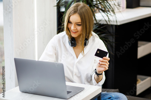 Young pretty girl spends time in a coffee shop. A young woman holds a black plastic card in her hands, and a laptop is next to her on the table