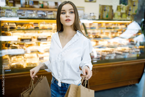 Young pretty girl spends time in a coffee shop. A young woman holds paper bags in her hands and stands near a shop window with sweets