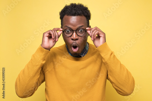 Young african american man in yellow sweater in shock, keeping mouth wide open, feeling stressed after having forgotten to pay bills in time. Studio shot