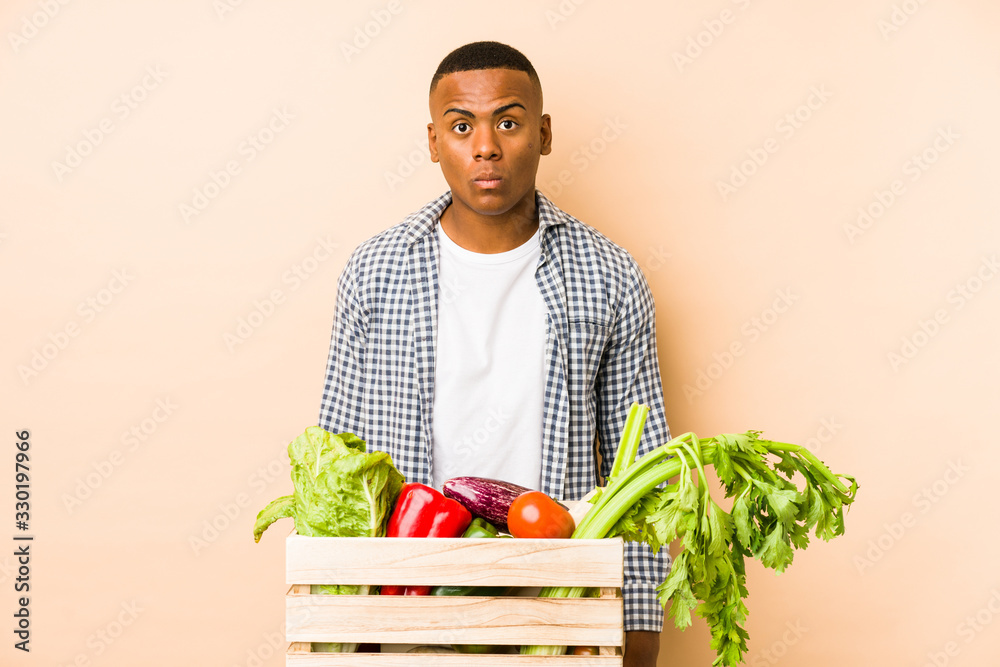Young farmer man isolated on a beige background shrugs shoulders and open eyes confused. Young farmer man isolated on a beige background shrugs shoulders and open eyes confused.