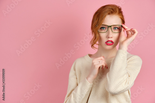 Beautiful redhead girl with red lipstick and manicure stands on a pink background and adjusting his glasses, looking at the camera photo