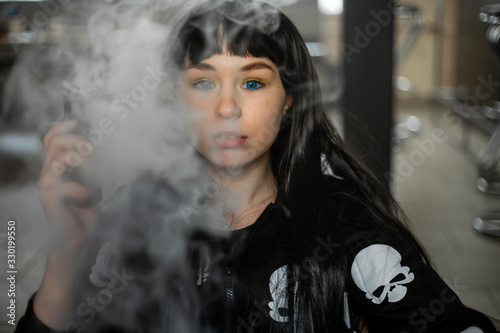 Vaping teenager. Young pretty white caucasian teenage brunette girl with blue eyes and problem skin smoking an electronic cigarette indoors. Deadly bad habit. Vape activity.