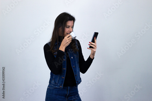 Portrait of attractive caucasian middle age woman model drinking coffee and looking at phone, isolated on gray background studio shot, black sweater, denim jacket, jeans, dark air. Place for your text