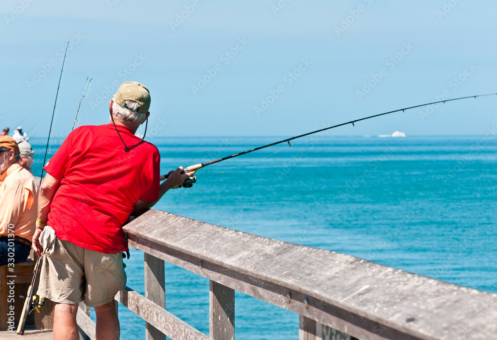 Back view, medium distance of a senior male in red shirt fishing off a wood pier in tropical, gulf of mexico on a sunny, winter day Back view, medium distance of a senior male in red shirt fishing off a wood pier in tropical, gulf of mexico on a sunny, winter day