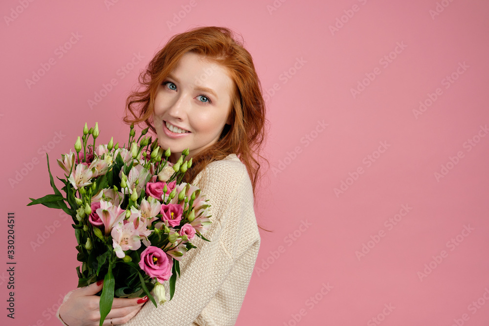 Beautiful red-haired girl hugs a bouquet with flowers on a pink background and gently smiles at the camera