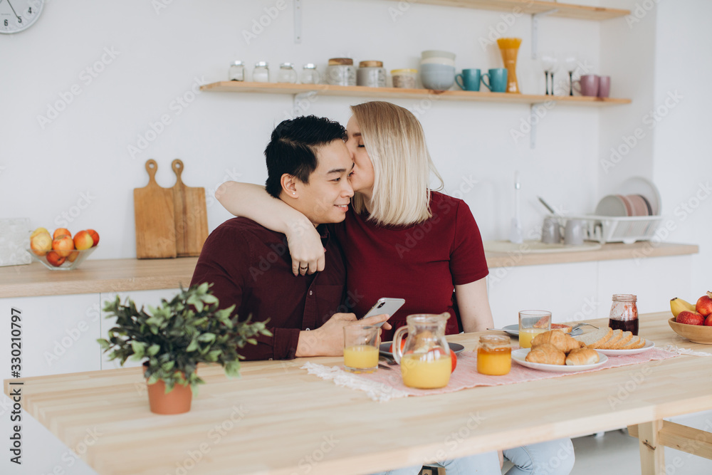Asian man and his caucasian blonde woman have breakfast. They spend time together in kitchen and look video on the smartphone.