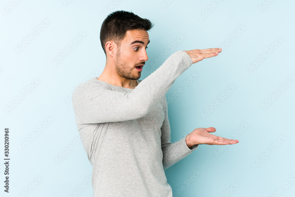 Young caucasian man isolated on blue background shocked and amazed holding a copy space between hands. Young caucasian man isolated on blue background shocked and amazed holding a copy space between hands.