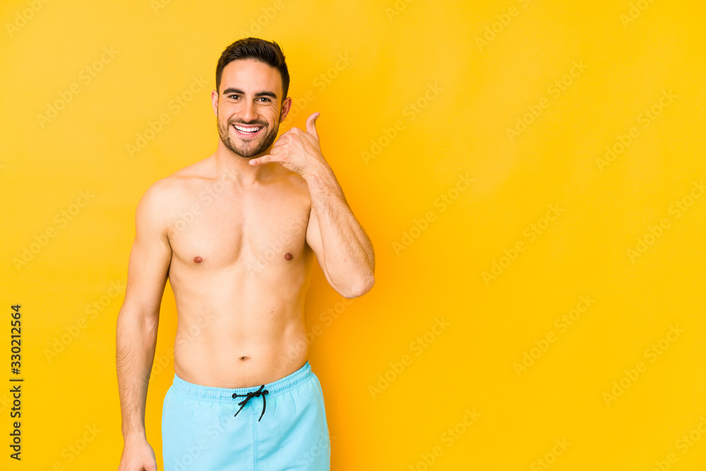 Young caucasian man with swimsuit isolated on yellow background Young caucasian man with trshowing a mobile phone call gesture with fingers.