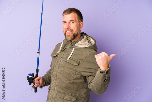Senior fisherman isolated on purple background points with thumb finger away, laughing and carefree.