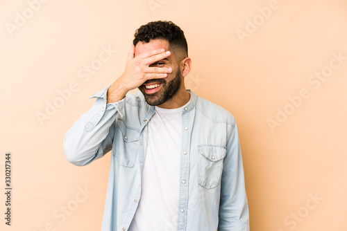 Young mixed race arabic man isolated blink at the camera through fingers, embarrassed covering face. © Asier