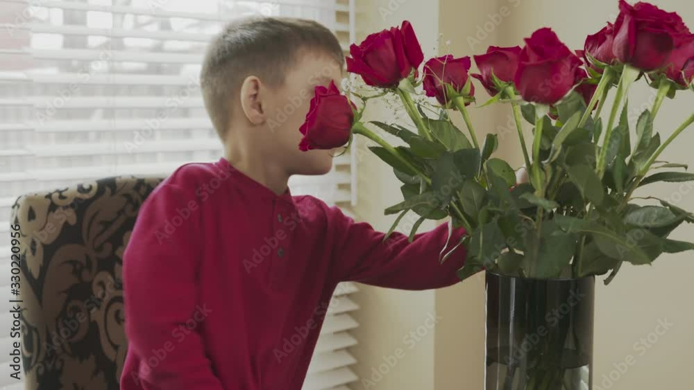 little happy child sniffs and enjoys the red roses that are in the vase on the table. Boy smiles and smells the flowers little happy child sniffs and enjoys the red roses that are in the vase on the table. Boy smiles and smells the flowers