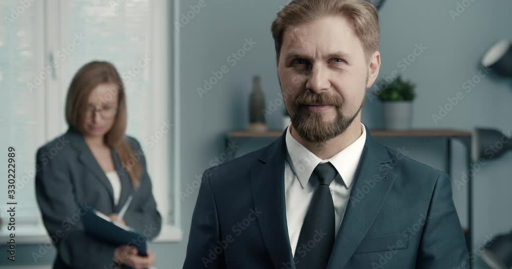 Mature bearded man in stylish suit smiling and looking at camera while his female colleague standing behind and reading documents. Two business partners working together at office Mature bearded man in stylish suit smiling and looking at camera while his female colleague standing behind and reading documents. Two business partners working together at office