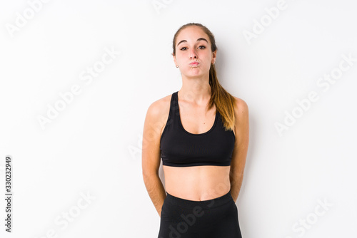 Young caucasian fitness woman posing in a white background blows cheeks, has tired expression. Facial expression concept.