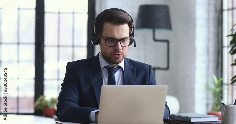 Confident businessman wears suit and headset makes conference video call on laptop. Male call center agent, customer support manager or telemarketer communicating in webcam chat on computer in office.