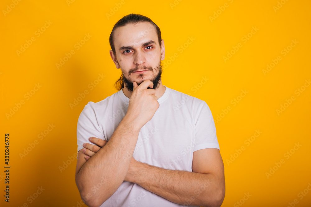 A guy with a beard in a white T-shirt on a yellow background in a thoughtful pose. A guy with a beard in a white T-shirt on a yellow background in a thoughtful pose.